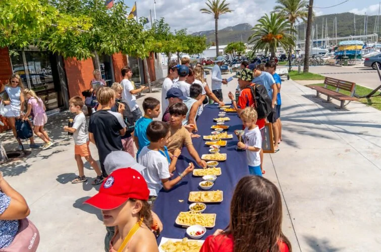 Niños y adultos en una fila disfrutando una comida al aire libre junto a un muelle con barcos, palmeras y montañas en el fondo.