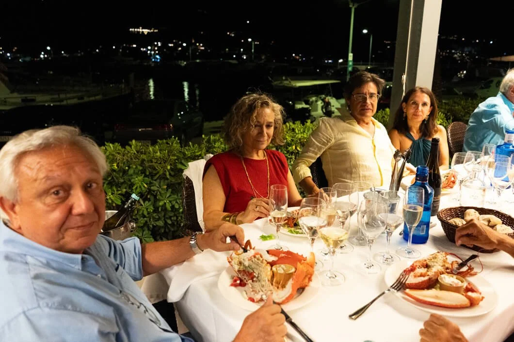 Grupo de personas sentadas en una mesa de cena al aire libre durante la noche, con comida y bebidas en la mesa, en un ambiente sofisticado con vista a un puerto o marina.