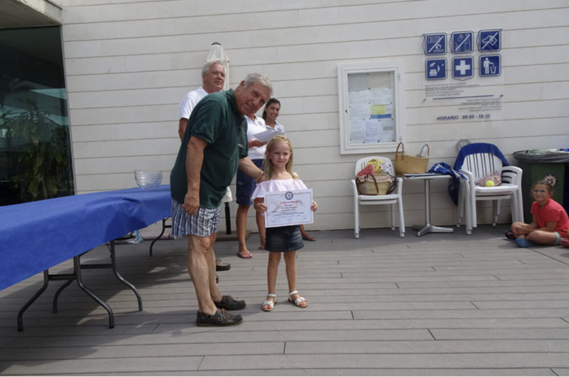 Niña recibe un certificado de reconocimiento de un hombre en un acto al aire libre.
