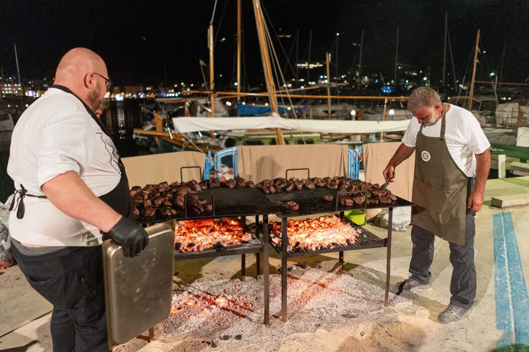 Dos chefs cocinan carne en una parrilla al aire libre en un puerto con barcos y mástiles visibles en el fondo en la noche.