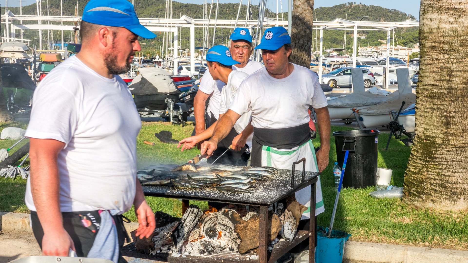 Hombres cocinando pescado a la parrilla al aire libre junto a un muelle con barcos y yates en el fondo, bajo palmeras.