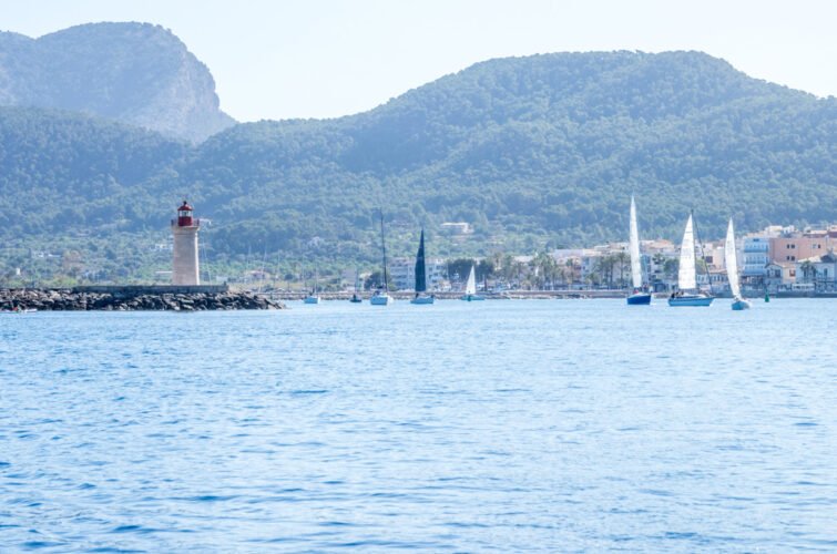 Vista de un mar con varios veleros navegando, un faro en la orilla y montañas verdes al fondo.