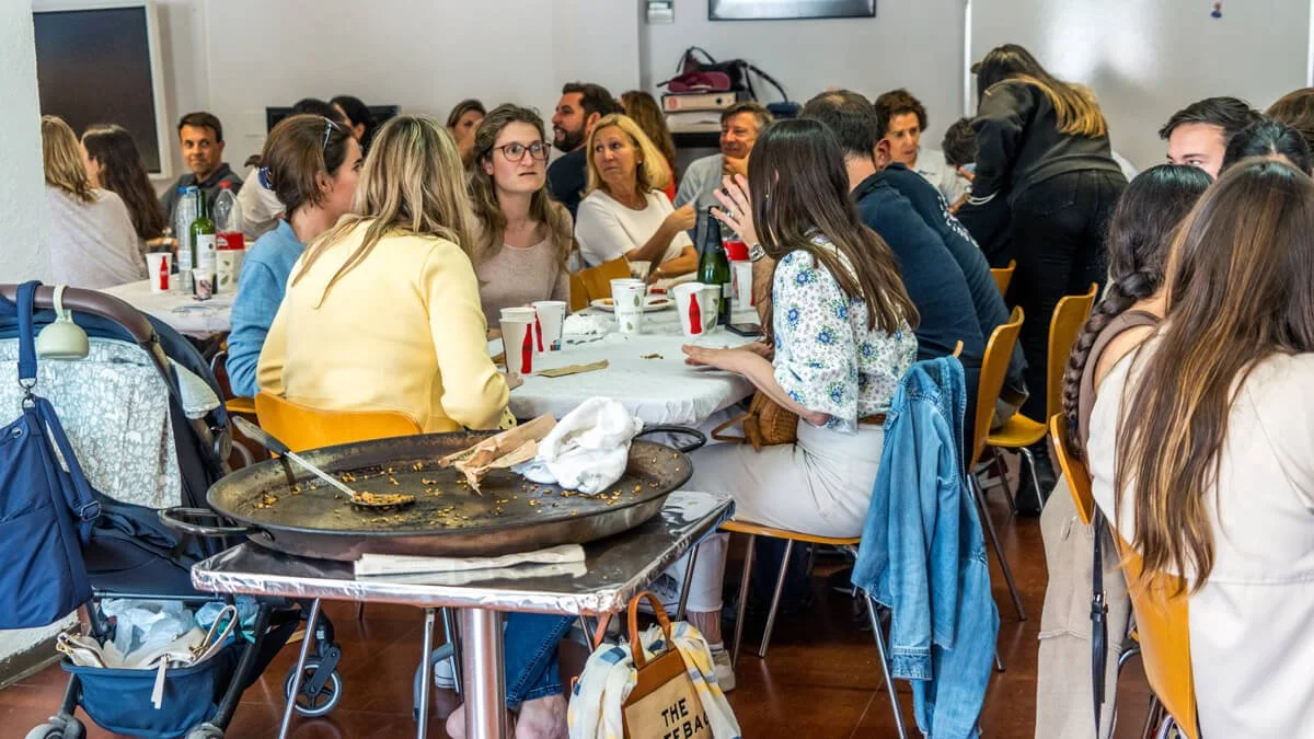 Grupo de personas sentadas en mesas durante una reunión o evento social en un ambiente interior, con comida y bebidas sobre las mesas.