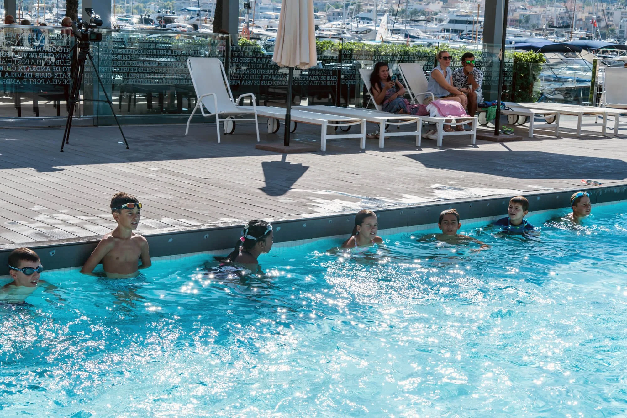 Personas en una piscina pública, con niños nadando y adultos observando desde la orilla, en un día soleado con vista a un puerto con muchos yates.
