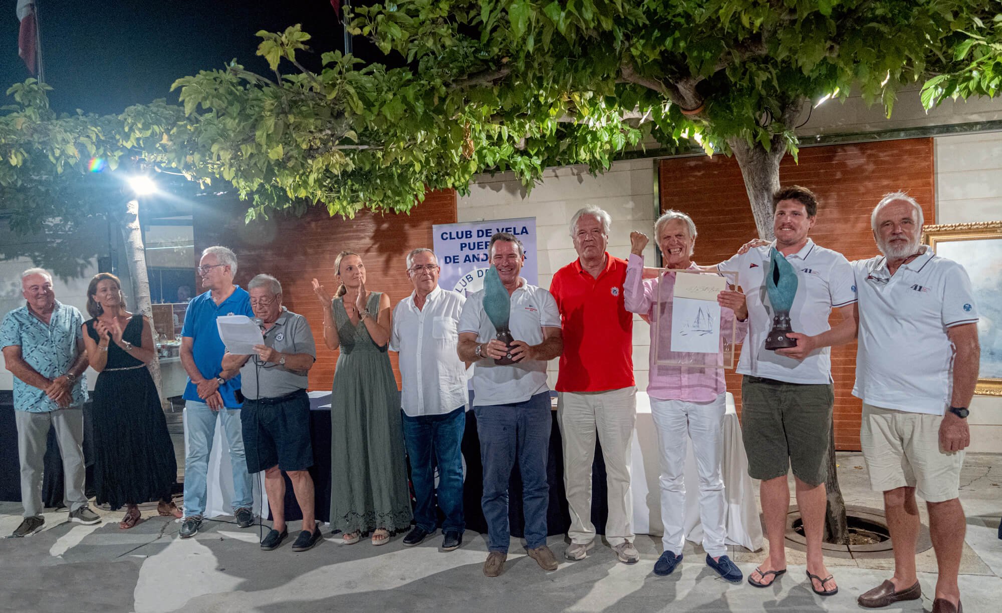 Grupo de personas celebrando en un evento en la calle bajo un árbol, algunos sosteniendo trofeos y un cuadro, con fondo de cartel que dice 'Club de Vela Puerta de And...'.