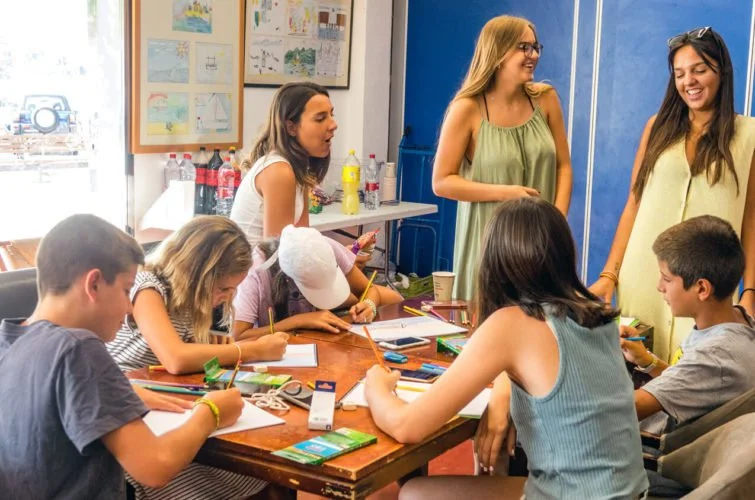 Niños y adolescentes en una mesa de clase, algunos dibujando y otros usando útiles escolares, mientras dos mujeres adultas conversan de pie al fondo.