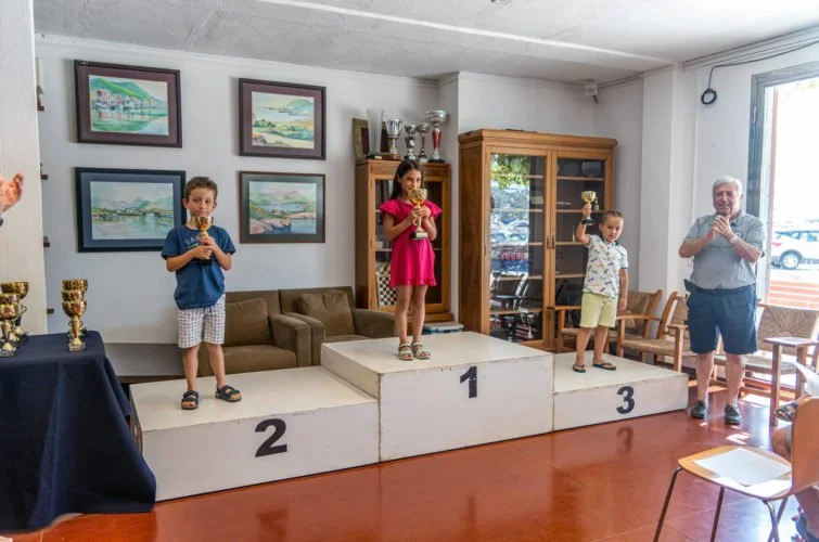 Niños en el podio de una competencia recibieron premios y un adulto a su lado en una ceremonia en un salón decorado con cuadros, trofeos y muebles de madera.