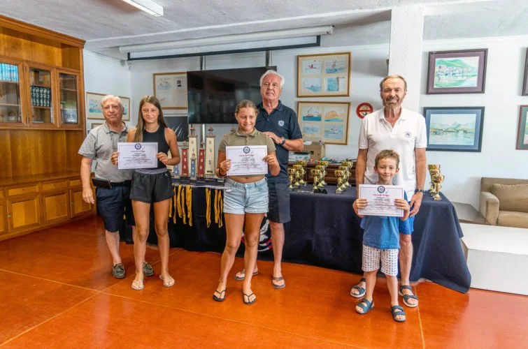 Grupo de personas en una ceremonia de premiación, con niñas sosteniendo diplomas y un hombre mayor. En el fondo, mesas con trofeos y decoraciones, en una sala con paredes con cuadros y estanterías de madera.