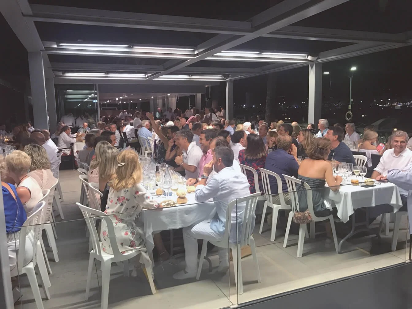 Imagen de una cena en un restaurante con muchas personas, mesas con blancos manteles y comida, en un ambiente moderno y elegante, de noche, con vista a la ciudad en el fondo.