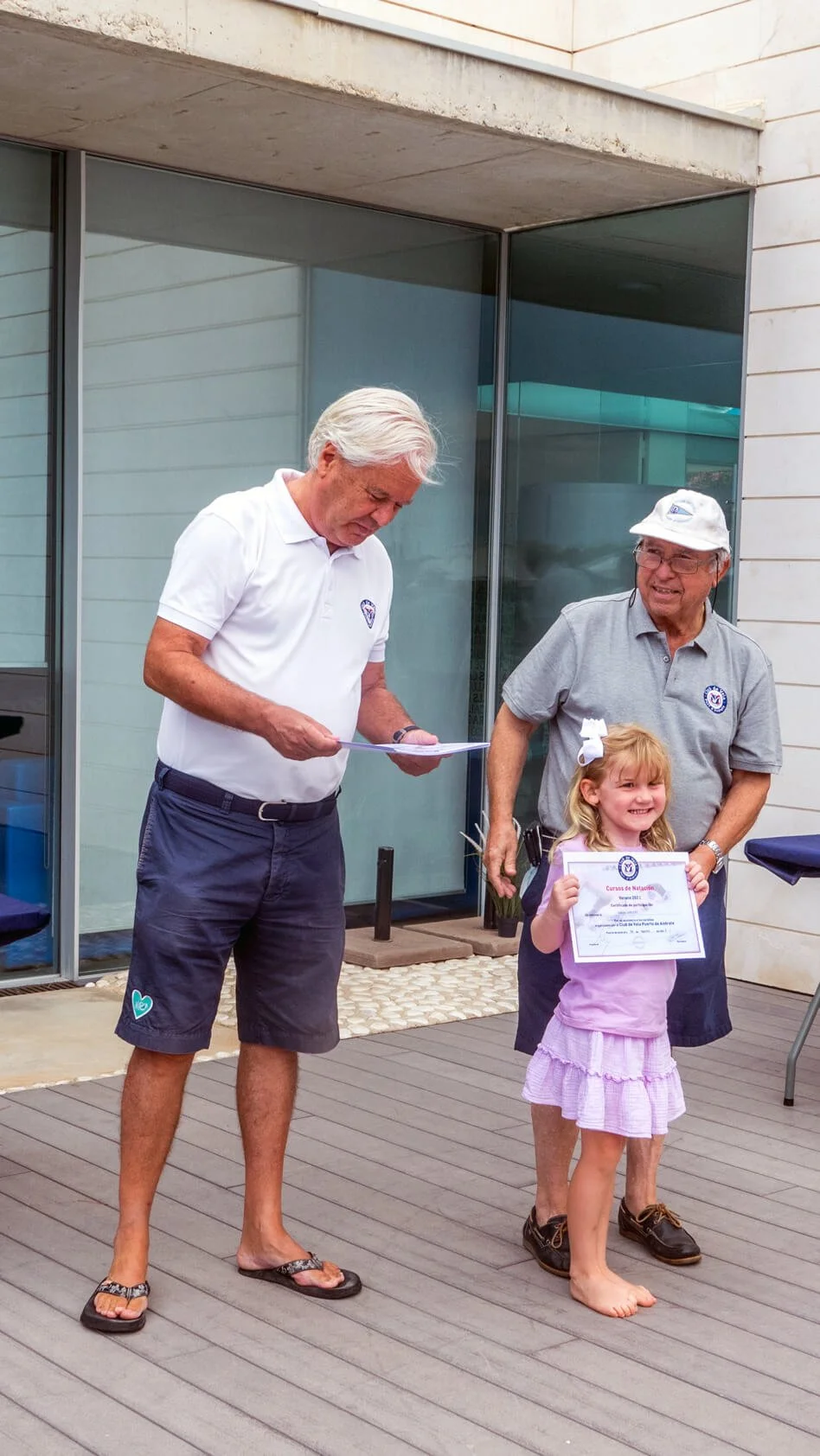 Una niña recibe un certificado de participación en una ceremonia en el exterior, rodeada de dos adultos. La niña sonríe, lleva un lazo blanco en el cabello y ropa rosa. Los adultos usan camisetas con logotipos y uno lleva gorra blanca, todos parecen 