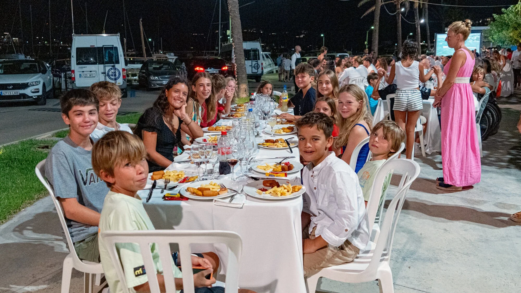 Grupo de niños y adolescentes sentados y de pie en una mesa larga con comida, en una cena al aire libre por la noche, con un fondo de autos estacionados y vegetación.