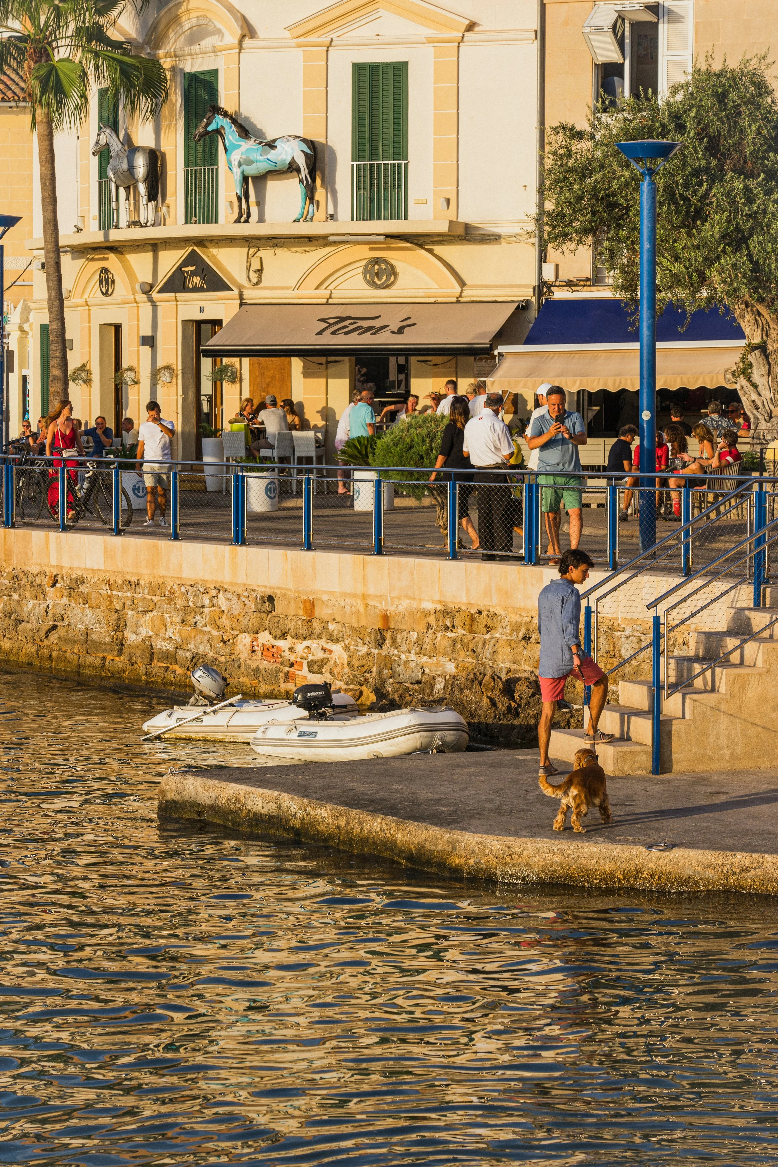Gente disfrutando en una terraza frente al mar, con perros, bicicletas y botes en el muelle, edificios de color amarillo y estatuas de caballos en la fachada.