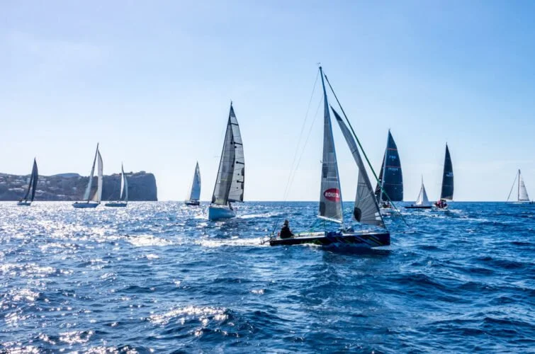 Varios barcos de vela navegando en el mar con un fondo de acantilados y cielo despejado.