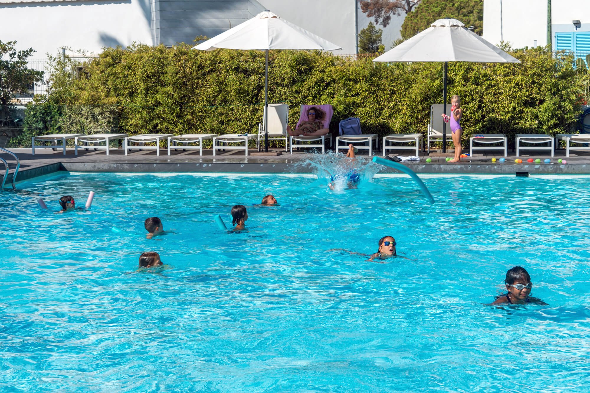 Niños y adultos en una piscina, algunos nadando y otros descansando en las sillas junto a la piscina, bajo sombrillas blancas, con arbustos verdes en el fondo y una estructura de construcción en el fondo.