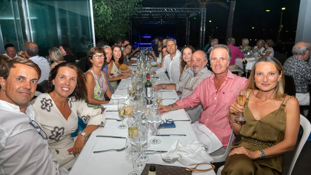 Grupo de personas en una comida o celebración en un restaurante o salón, sentadas a una mesa larga con comida y bebidas, en un ambiente nocturno.