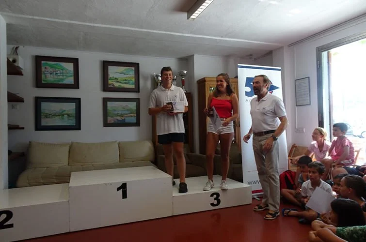 Persona en un podio de premiación con niños y adultos en una sala decorada con cuadros y ventana grande.