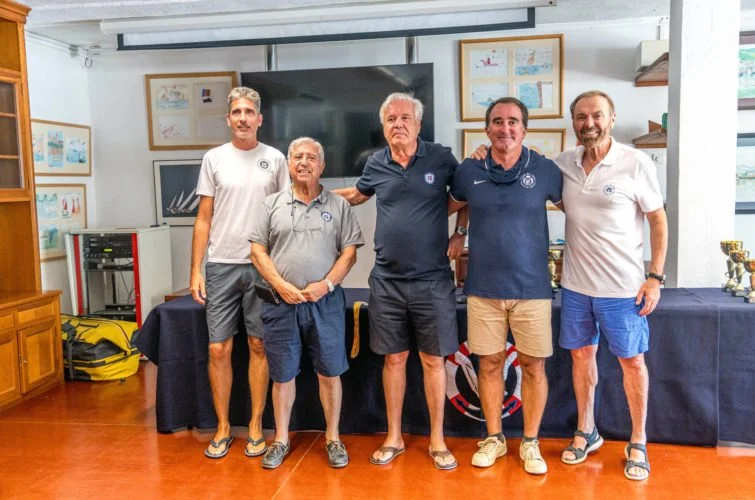 Cinco hombres en una sala, algunos con camisetas de fútbol, posando juntos en frente de una mesa con trofeos, en un evento social o deportivo.