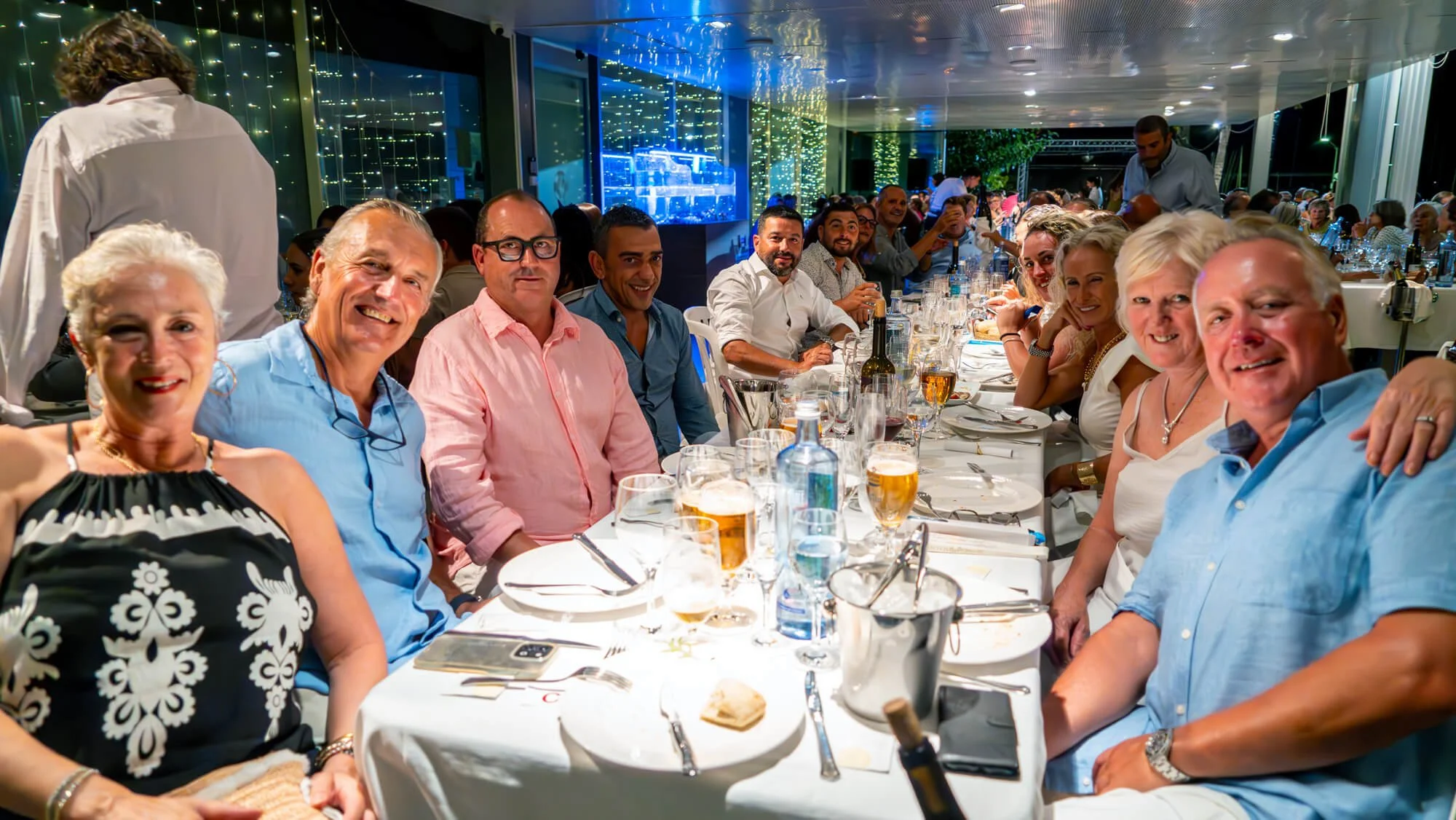 Grupo de personas sentadas en una mesa en un evento social, usando ropa de vestir, con copas de cerveza y agua, en un ambiente iluminado y decorado con luces