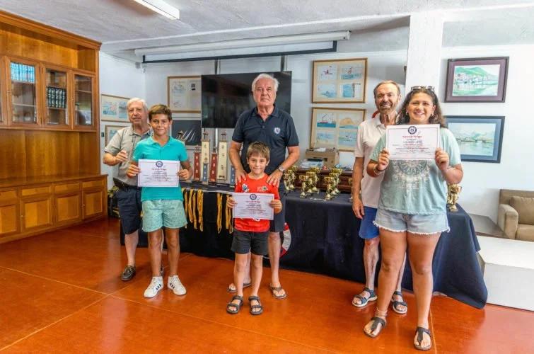 Cinco personas en una sala de premiación, tres niños y dos adultos, sosteniendo certificados y trofeos, en un evento de reconocimiento académico o deportivo.