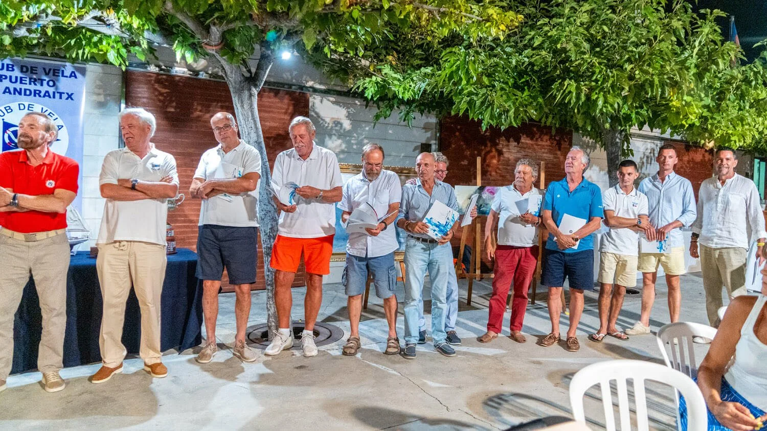 Grupo de personas en una ceremonia al aire libre, algunas leyendo y otras con libros en las manos, con un árbol y decoraciones en el fondo.