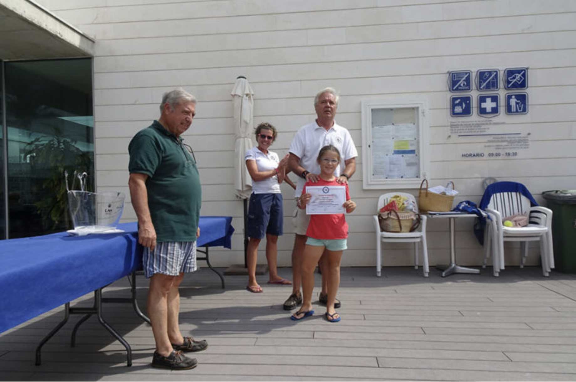 Grupo de personas en un evento de reconocimiento, una niña recibe un certificado, fondo con señalización de servicios médicos y un espacio de descanso con sillas y bolsas.