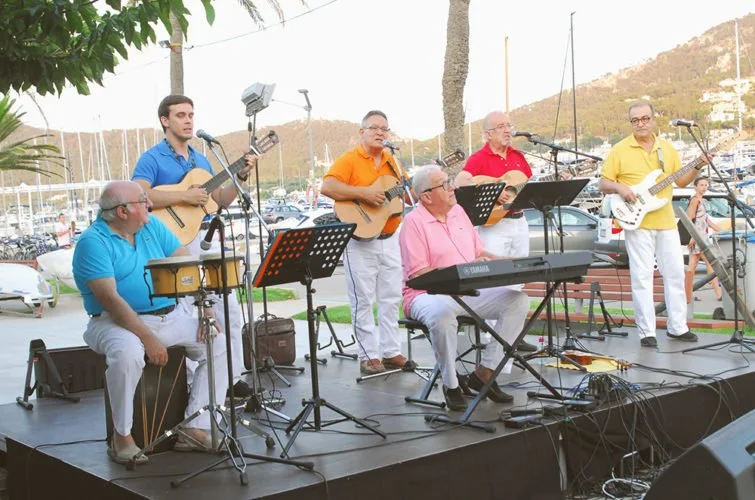 Grupo de seis músicos tocando en un escenario al aire libre con instrumentos acústicos y eléctrico en un puerto con barcos y montañas de fondo.