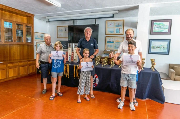 Niños y adultos en una ceremonia de premiación en una sala con estanterías de madera. Los niños sostienen certificados y uno de los adultos muestra un pulgar arriba.