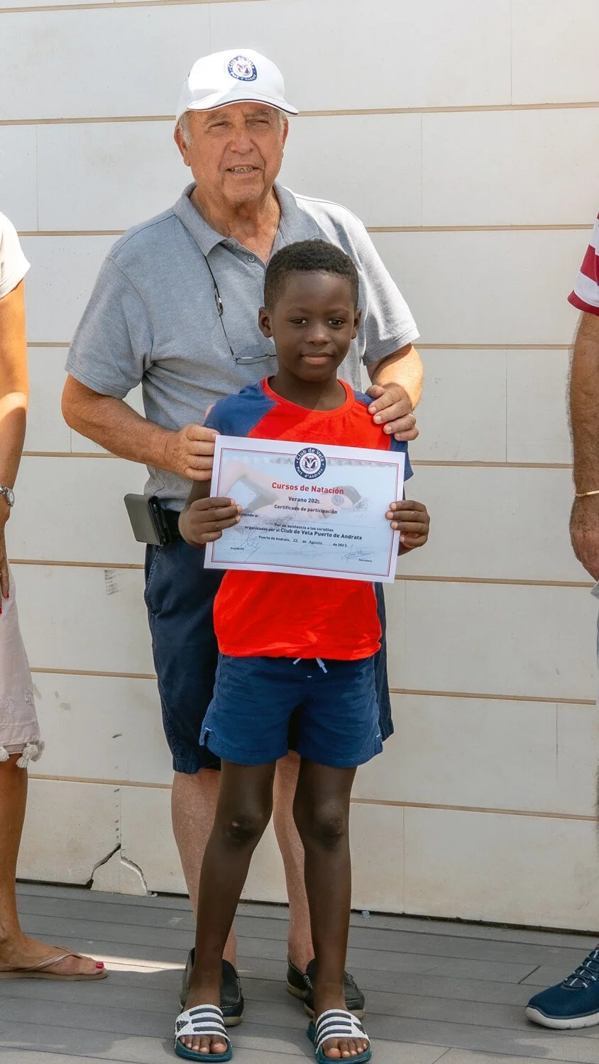 Un niño sostiene un certificado de participación en un curso de natación, acompañado por un adulto mayores, en un evento al aire libre.