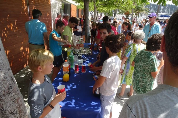 Muchas personas en una fiesta al aire libre con mesas de comida y bebidas, árboles y un edificio de fondo.