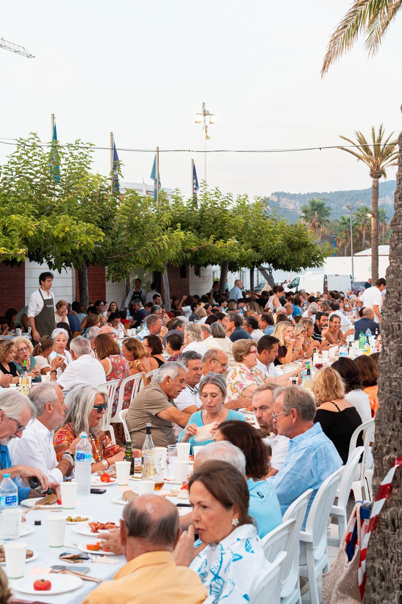 Una gran reunión de personas sentadas en mesas al aire libre, disfrutando de una comida en un ambiente festivo con palmeras y banderas en el fondo.