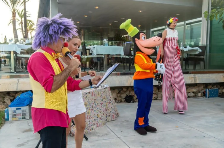 Un hombre disfrazado de payaso y un personaje de Mickey Mouse ofrecen un espectáculo para niños frente a un restaurante, con una mujer que actúa de payaso en el escenario.