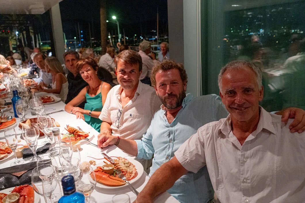 Grupo de personas cenando en un restaurante con vista a un puerto, alrededor de una mesa con comida y bebida, durante la noche.