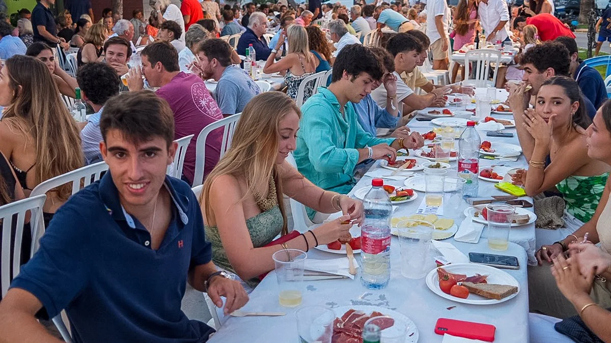 Grupo de personas jóvenes sentadas en una mesa larga al aire libre, comiendo y bebiendo durante un evento social.
