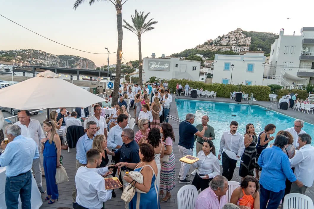 Reunión social junto a una piscina en un área exterior con muchas personas, mesas y sillas, palmeras y un paisaje montañoso con casas.