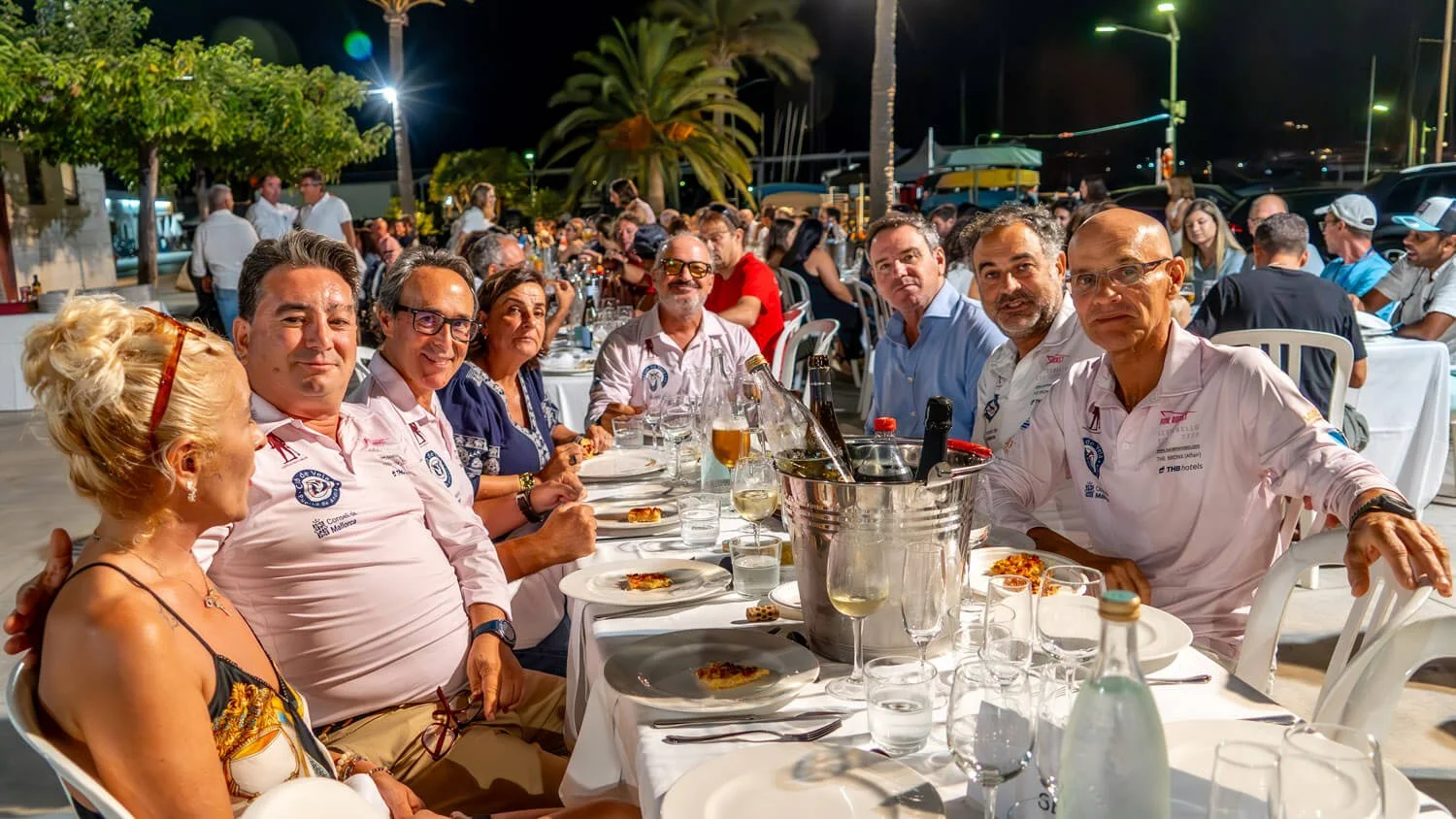 Grupo de personas en una cena al aire libre por la noche, en una mesa con comida y bebidas, en un ambiente festivo y social.