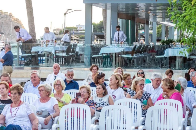 Grupo de personas mayores sentado al aire libre en un evento social, con una estructura moderna de fondo con mesas y sillas