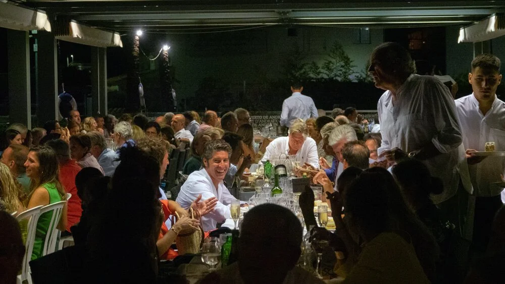 Personas en una cena en un restaurante por la noche, charlando y sonriendo, con mesas llenas de comida y bebida.