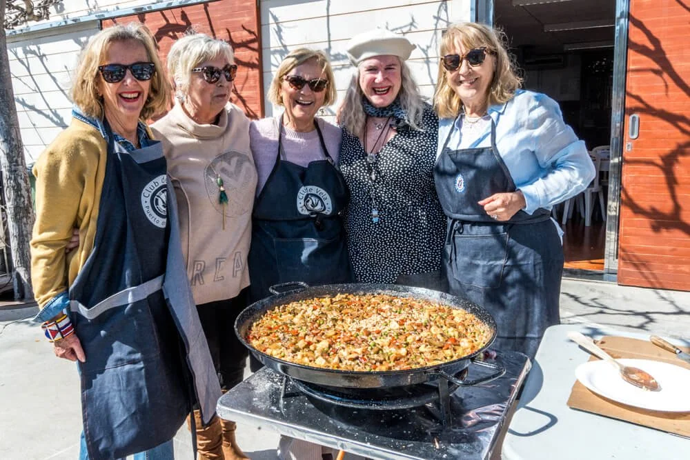 Cinco mujeres mayores son sonriendo y posando con una gran paella en una mesa al aire libre. Todas llevan gafas de sol y delantales, y parecen disfrutar de una reunión social.