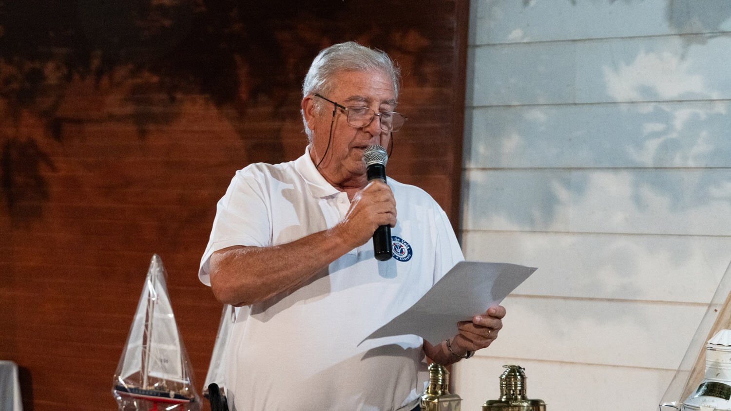 Hombre mayor con gafas y camiseta blanca leyendo un papel y usando un micrófono en un ambiente interior.