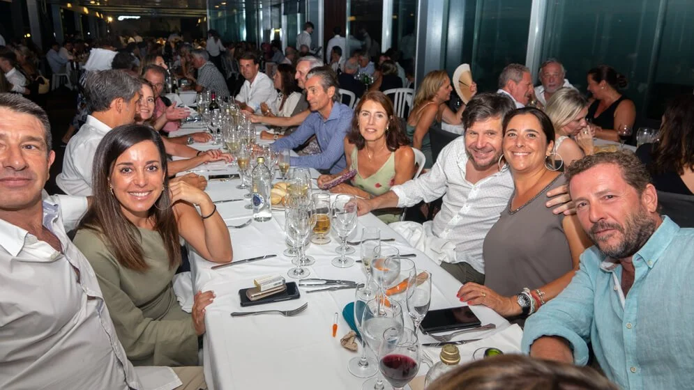 Grupo de personas en una cena formal en un restaurante, sonriendo y disfrutando de la compañía, con una larga mesa y decoraciones elegantes.