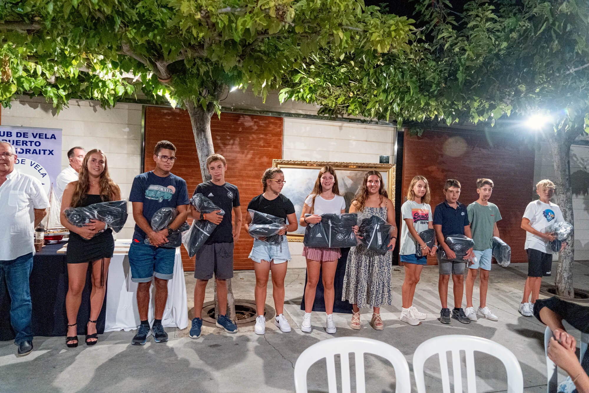 Grupo de personas, incluyendo niños y adultos, en un evento al aire libre por la noche, con algunos sosteniendo bolsas negras. Hay un árbol y una pared con un cuadro en el fondo, y una mesa con premios o regalos.