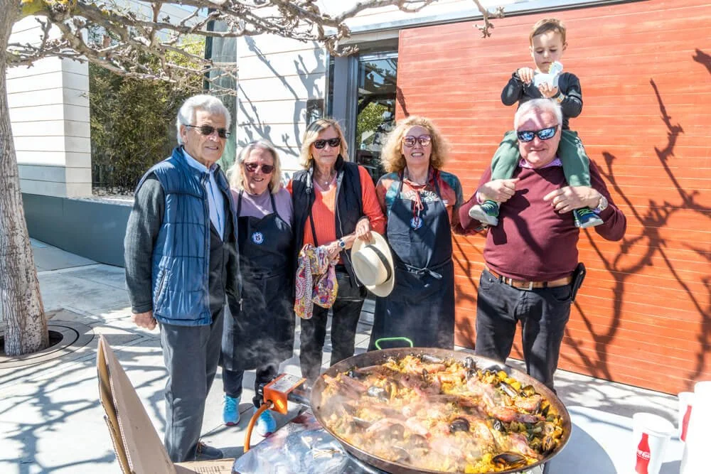Grupo de seis personas disfrutando de una comida al aire libre, con una gran paella en una mesa. Un niño está en los hombros de un hombre. Todos parecen estar en un día soleado, en un ambiente casual y alegre.