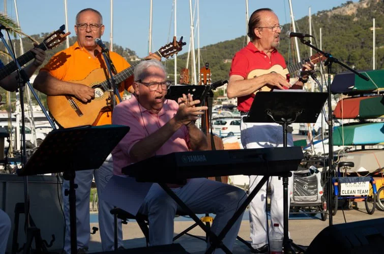 Tres músicos tocando instrumentos en un escenario al aire libre cerca del puerto, con montañas y yates de fondo.