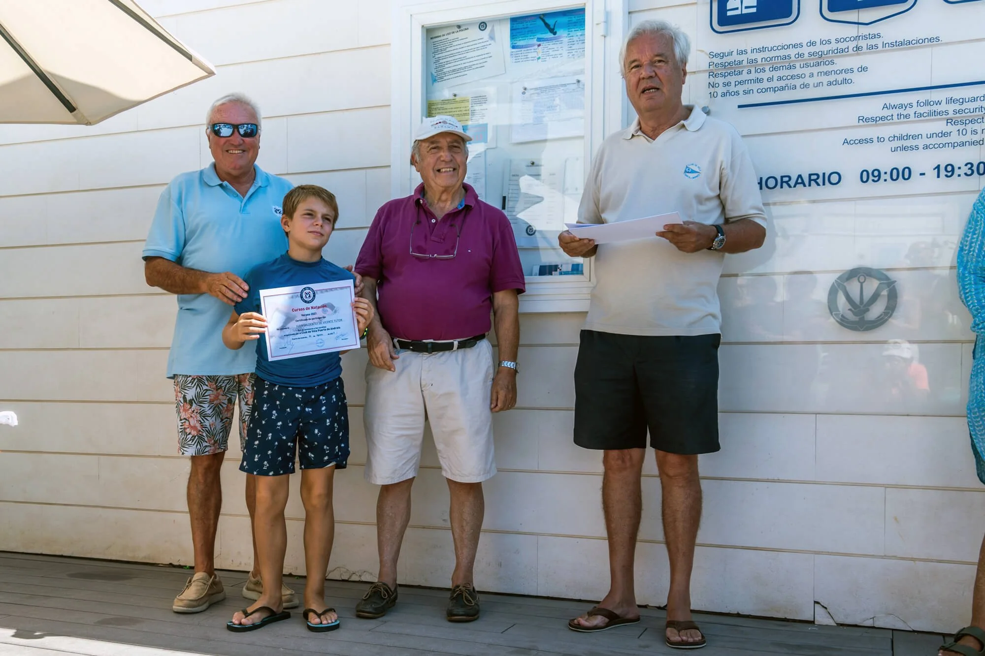 Grupo de personas de diferentes edades posando en un edificio con información en la pared. Un niño sostiene un certificado de natación. Todos parecen estar felices después de una ceremonia o reconocimiento.