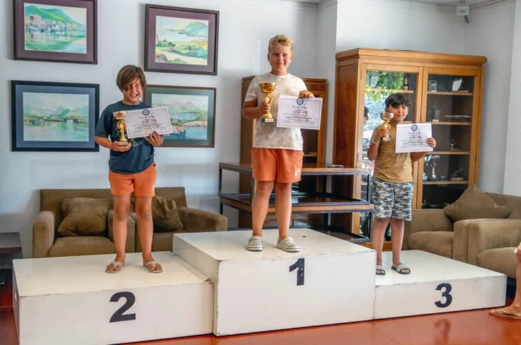 Tres niños en un podio de ganadores en una ceremonia de premiación, con trofeos y certificados, en una sala con cuadros en la pared y sofá.