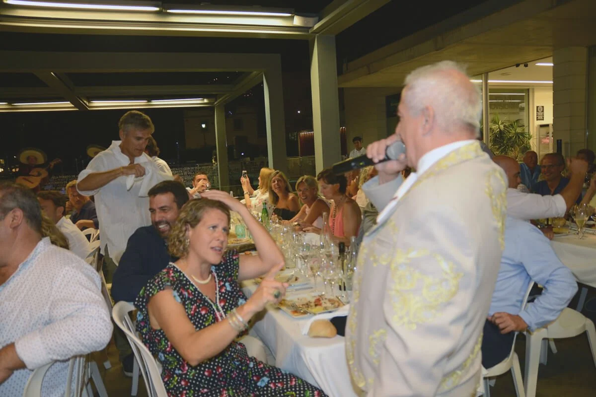 Hombre mayor con micrófono en mano en una fiesta o cena con gente sonriendo y disfrutando en una mesa larga