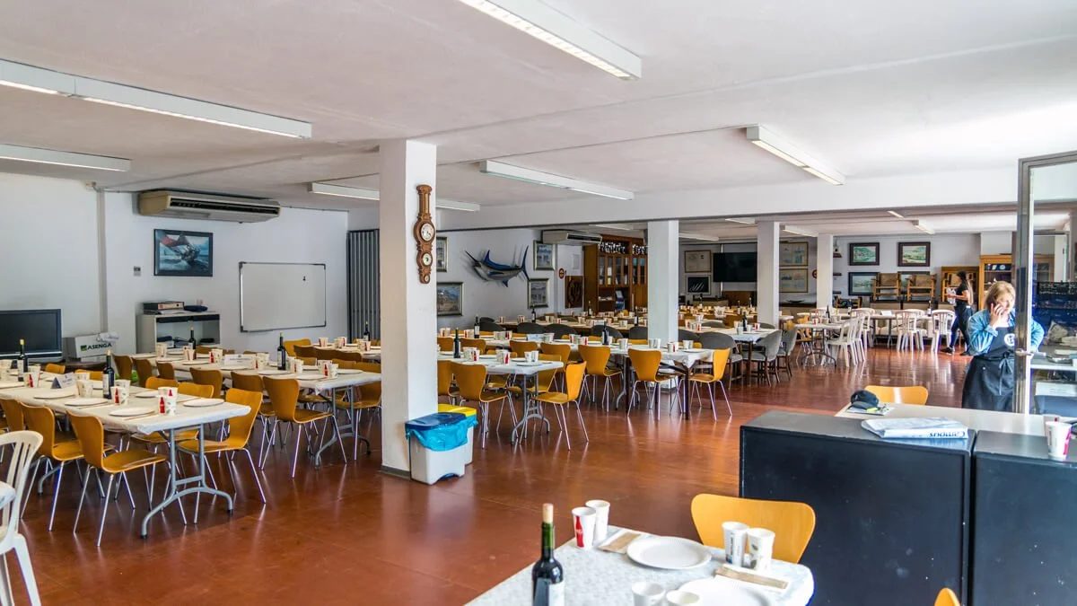 Un comedor con mesas y sillas amarillas y blancas, preparadas para cenas, en un ambiente amplio con paredes blancas, cuadros, y una joven atendiendo.