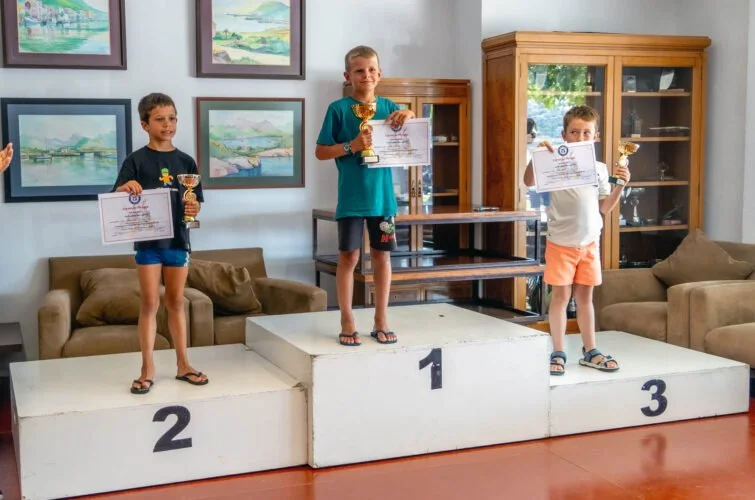 Tres niños en un podio de premios, sosteniendo trofeos y diplomas, celebrando una competencia en un salón con pinturas y muebles de madera.