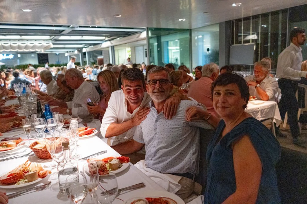Personas en una cena elegante en un restaurante, con comida y bebida en la mesa, muchas personas en el fondo y un ambiente formal.