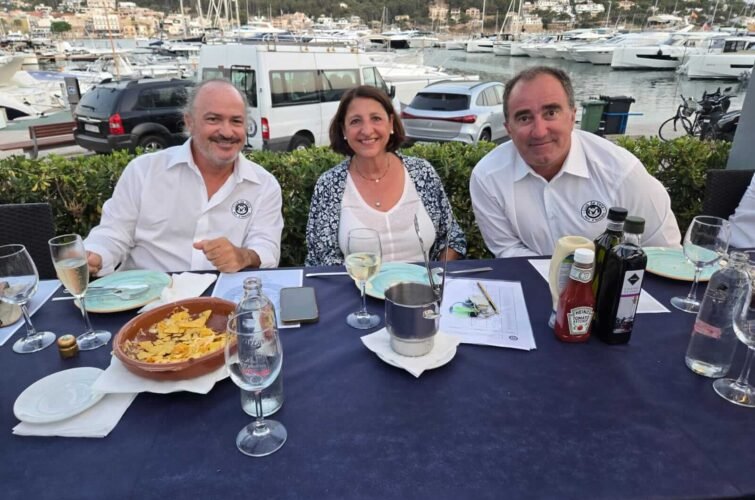 Tres personas sentadas en una mesa al aire libre con un muelle y yates de fondo, comiendo y bebiendo en un restaurante frente al mar.